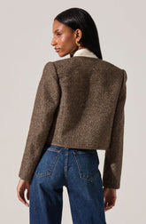 A woman is wearing a brown, textured, cropped blazer with slightly padded shoulders, paired with dark blue high-waisted jeans. The blazer has a subtle herringbone pattern and a structured fit. She also has on a white shirt underneath the blazer and gold earrings.